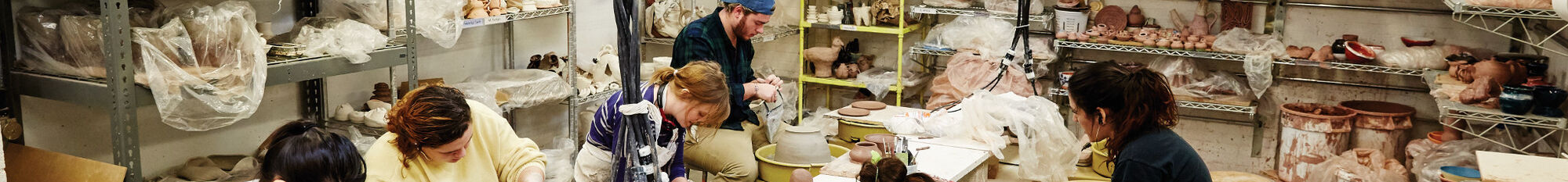 A wide shot of a ceramics studio, featuring students working with pottery wheels and other tools.