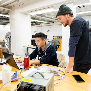 Two students look at a computer on a desk in an industrial work space.