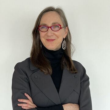 Shawn Smith wears a grey blazer with a black turtleneck layered underneath, red eye glasses and silver earrings. Her arms are folded across her chest and she is smiling at the camera.