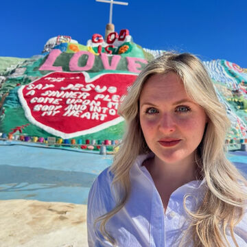 Annalise has long blonde hair and she wears a white button-down shirt and stands in front of Salvation Mountain.
