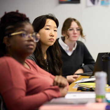 Three students in a classroom sitting a desk listening to someone speak.