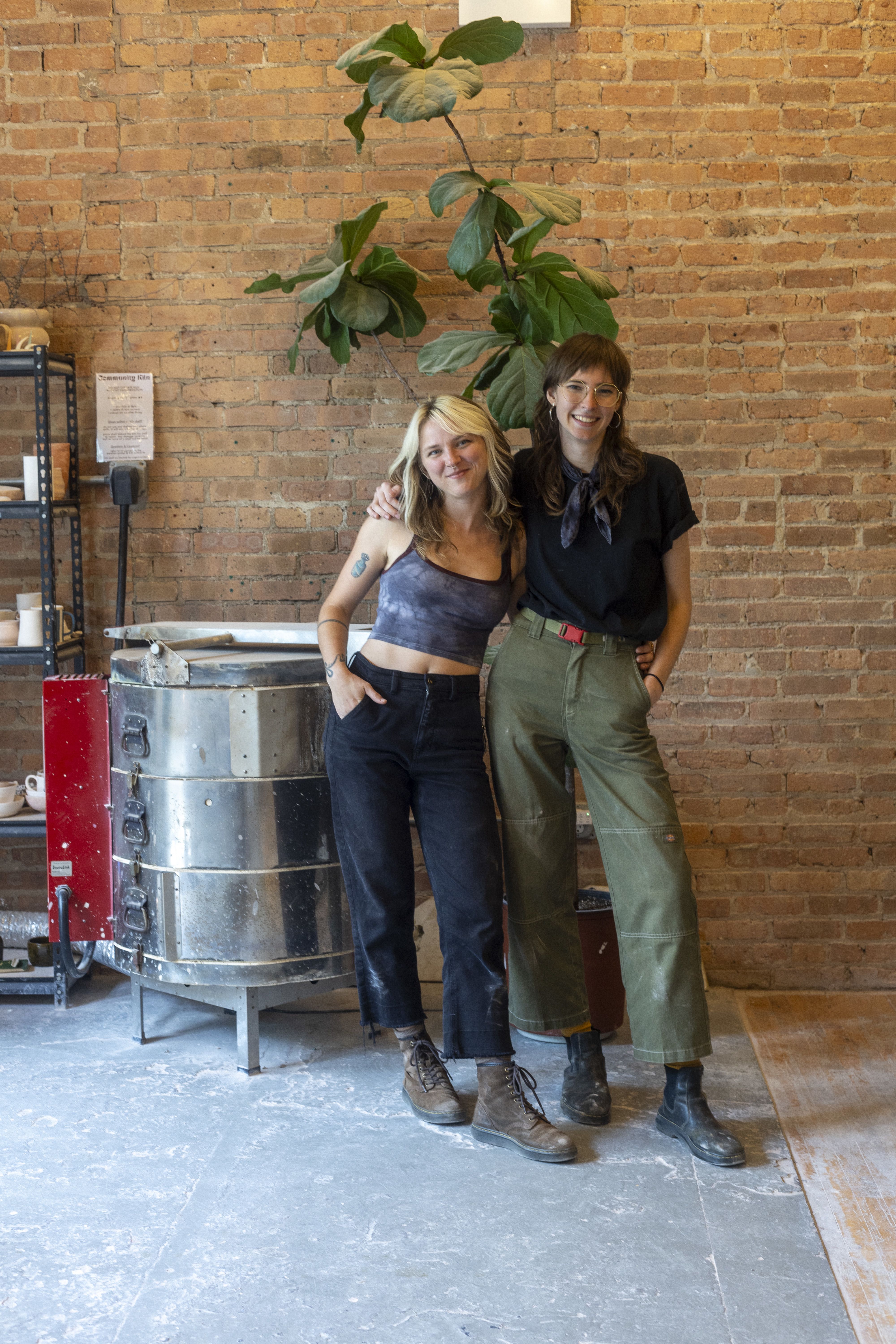 Two artists pose in front of a brick wall and kiln