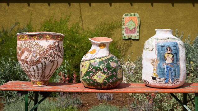 Four ceramic artworks are in view in green garden-like surroundings. Three of which are in the foreground on a rough orange rectangular table. The other artwork is a ceramic painting in the background hanging on an outdoor wall. 