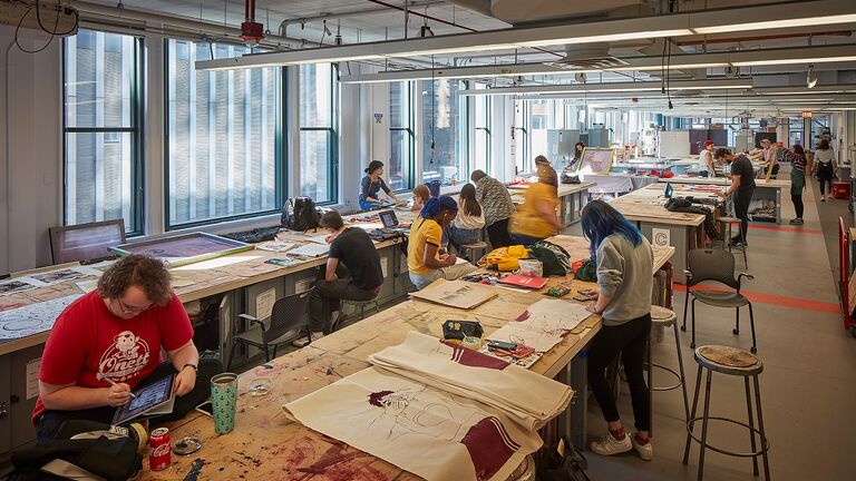 Students working individually in a sprawling studio space with large windows showcasing downtown Chicago architecture.