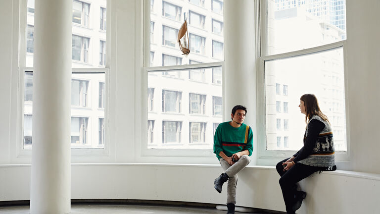 Two people sitting in a space with tall windows. 