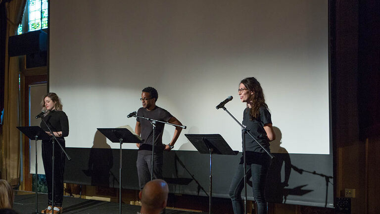 Three people reading out loud into a microphone from works placed on a music stand in front of them. 