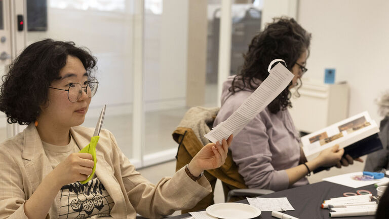 Two people sitting at a desk. One person is cutting paper with scissors, the other person is reading a book. 