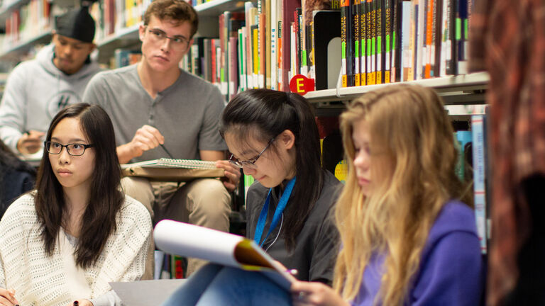 5+ students sitting in a library, some of them reading