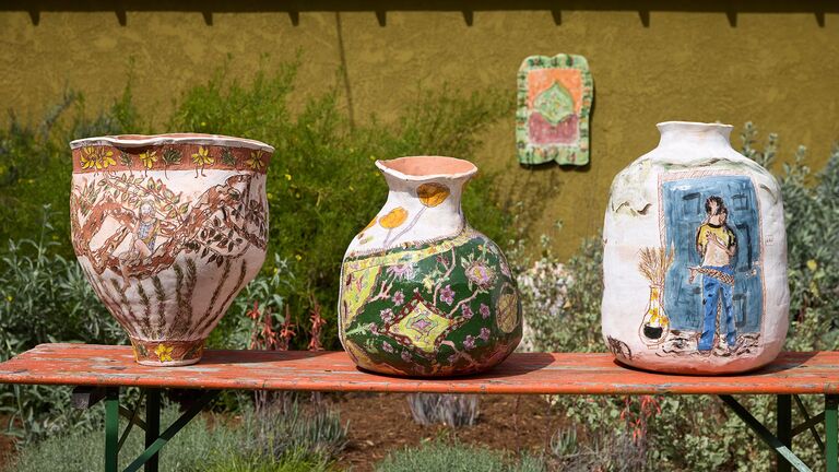 Four ceramic artworks are in view in green garden-like surroundings. Three of which are in the foreground on a rough orange rectangular table. The other artwork is a ceramic painting in the background hanging on an outdoor wall. 