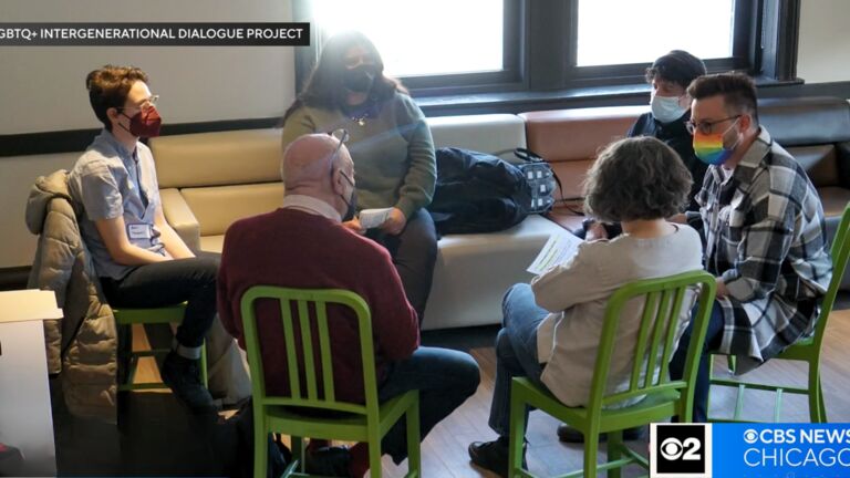 A group of people engages in a discussion while seated in a circle at the LGBTQ+ Intergenerational Dialogue Project event. They are indoors, wearing masks, and seated on green chairs and a couch.