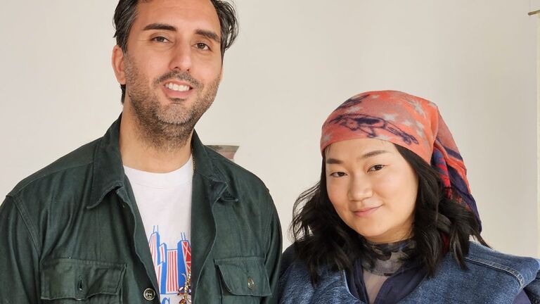 A man and woman stand side by side, smiling. The man wears a green button-up shirt over a graphic T-shirt, while the woman wears a denim vest, a long sleeve shirt, and a patterned headscarf.