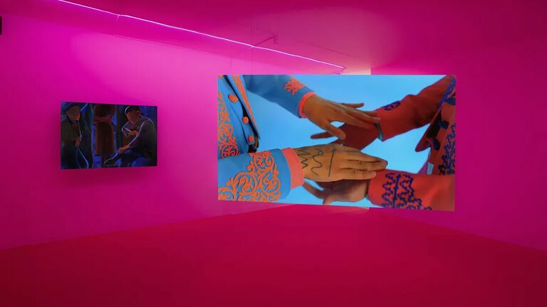 An art installation featuring two vibrant screens, one showing hands in colorful attire, the other depicting people sitting. The walls and floor are vividly lit in pink with neon lights overhead.