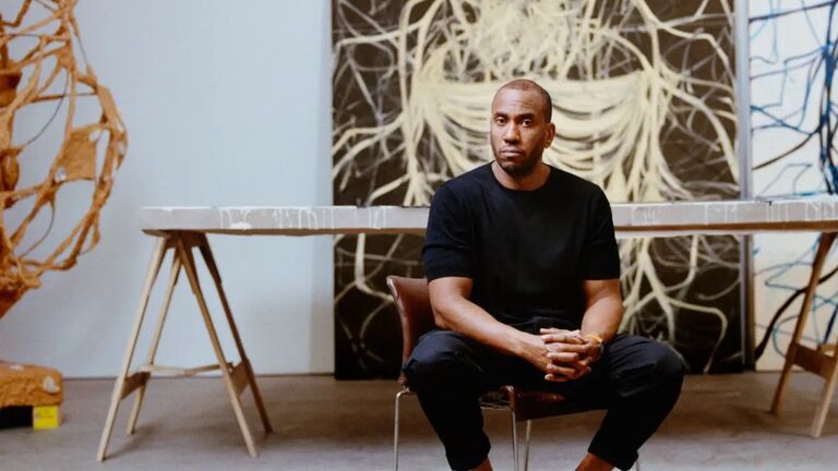 Rashid Johnson sits on a chair in a gray minimalist industrial studio space with his paintings and sculptures behind him