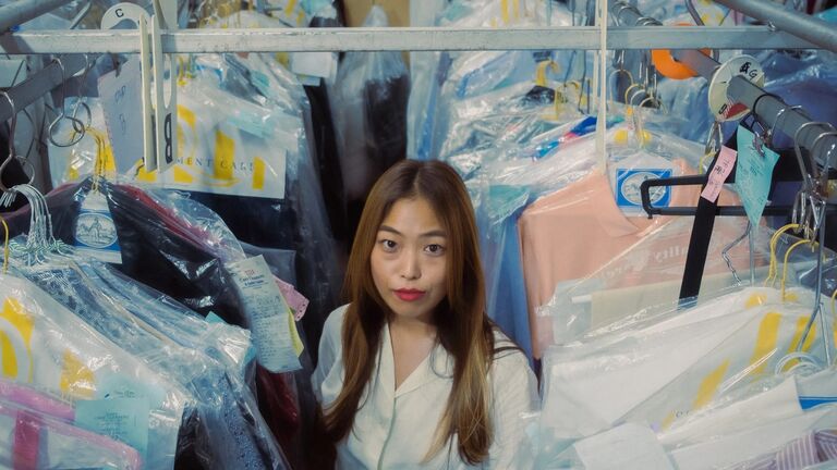 Artist Katie Chung stands amid hanging rows of plastic-covered clothes at a dry cleaners. She looks up at the camera. She wears red lipstick and a white shirt and has long reddish brown hair.