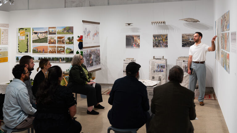 An adult gesturing to mounted wall art to a room full of seated adults in a gallery space