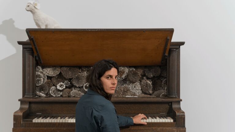 A person sits at an upright piano, looking at the camera. The piano lid is open, revealing honeycombs instead of strings. A white cat figurine is placed on top of the piano.