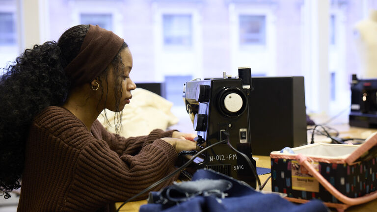 An adult person with a deep skin tone and curly black hair works with a sewing machine.