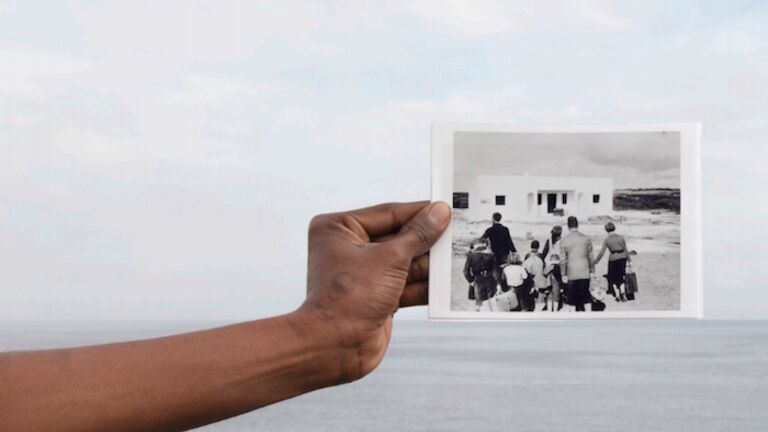 A hand holds up a black and white photo of a family with suitcases arriving at a white house before a vista of open water