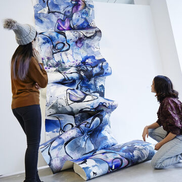 Two students looking at a long piece of canvas vertically cascading down a wall covered in blues an purples and black swirls in watercolor.