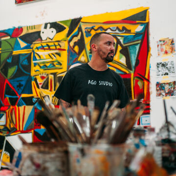 An artist stands in front of a colorful work of art in his studio