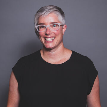 Sarah Ross smiles at the camera in a black tee shirt. Her salt and pepper hair is cut short and she wears glasses.