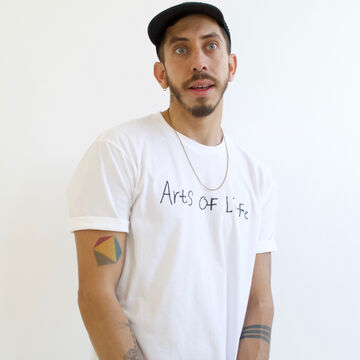 Vincent Uribe stands against a white background wearing a white t-shirt that reads Arts of Life and a black baseball cap.