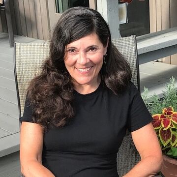 Cathy sits in a patio chair. She wears a short sleeve, black dress and she has dark, long hair.