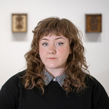 Maddie faces the camera wearing a black sweater with a black and white check collar. She has curly red hair and bangs. 