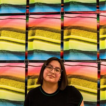 Marylu wears a black short sleeve shirt and stands in front of a wall of multicolor screens.