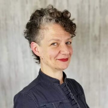 Rachel stands before a grey background wearing a navy jacket that has a ruffled placket.