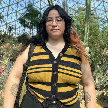 Sarita wears a black and yellow strip vest and stands inside a biodome.