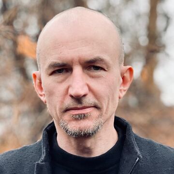 Lukasz wears a black jacket and shirt while standing in front of a leafless tree.