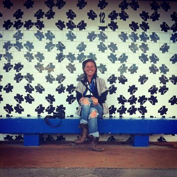 Meredith sits on a royal blue bench in front of a tiled wall. She wears jeans with holed in the knees, cuffed above brown boots.