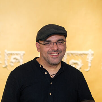 Shaurya wears a black newsboy cap and a black shirt with buttons.