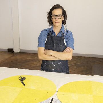 Diana stands, arms crossed, wearing a short sleeve denim shirt beneath a dark denim apron.