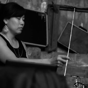 Black and white image of an Asian woman giving a sound performance and holding a thin metal rod with her eyes closed. 