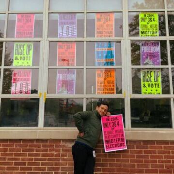 An arts administrator poses with a poster in front of a window full of posters