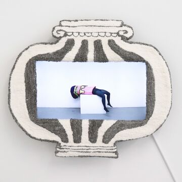 Image of a video screen framed by a tufted fiber column condensed into vessel shape. The video depicts a performer laying on their back on a pedestal and wearing a tufted column costume.