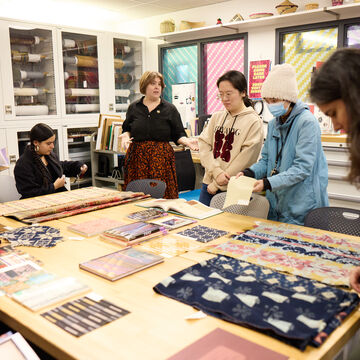 A photo of the Textile Resource Center including students and professors overlooking a table of selected books and textile objects.