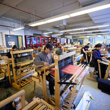 a photo of a weaving studio with rows of floor looms and student weaving various projects.