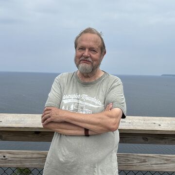 George Liebert, an elder person with a fair skin tone and short gray hair with a beard, standing in front of a body of water with crossed arms.