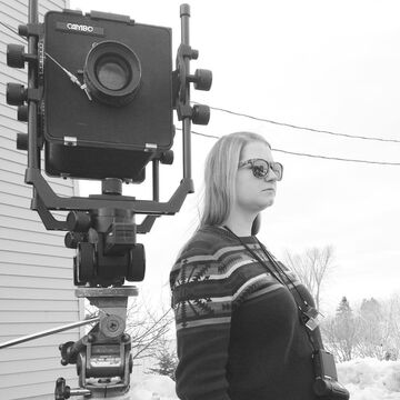 A black and white photo of Meghan Rose, SAIC faculty member, standing next to a camera.
