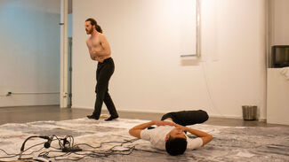 A white man standing and an Asian man lying on an art installation in a gallery space and performing 