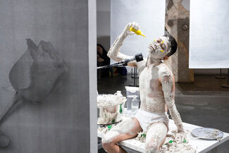 Performance art piece featuring an Asian person covered in a pale, clay-like substance and squirting a bottle of yellow paint on their face. 