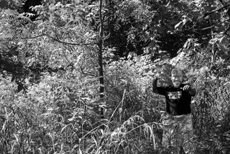 Black and white photo by Cecil McDonald, Jr. of a masked subject gesturing with their arms in a wooded area. 