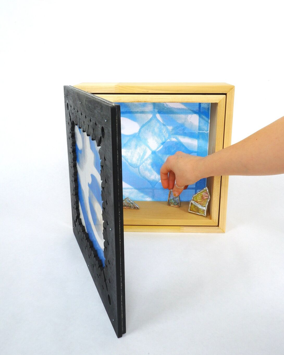 A hand is reaching into a wooden box with an open front panel, adjusting small objects inside. The box has a blue patterned background and a black frame.