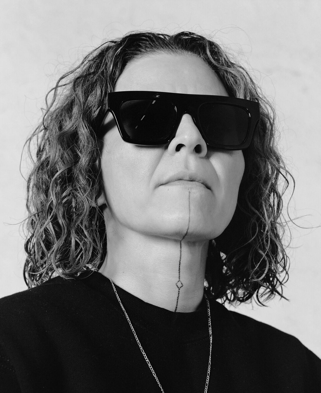 A person with shoulder length curly hair and sunglasses on, black shirt and chain necklace. Black and white photo