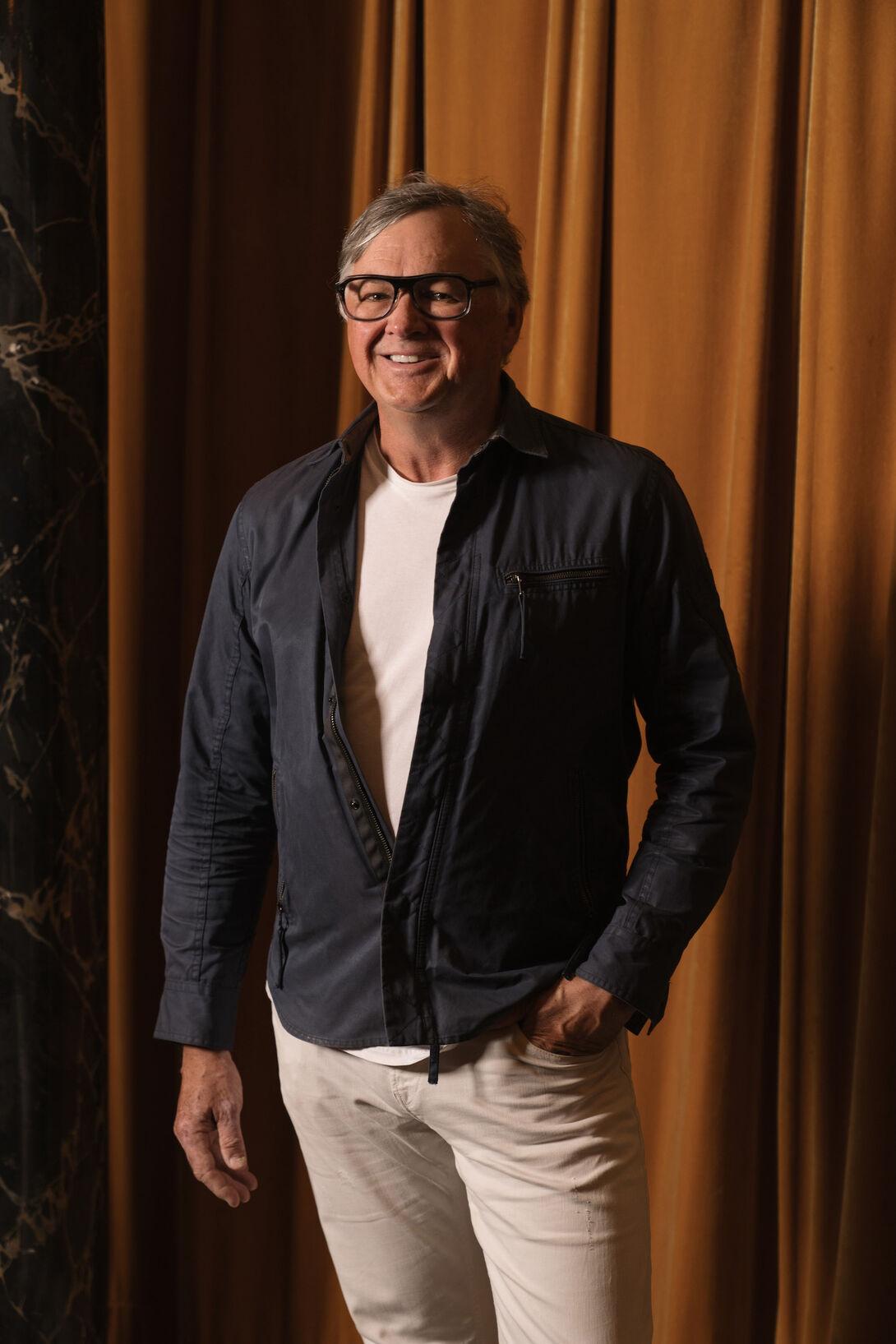 A man with gray hair and glasses and a black button down partially open over a white t-shirt, khakis. He has a hand in one pocket and is standing against a mustard colored fabric background