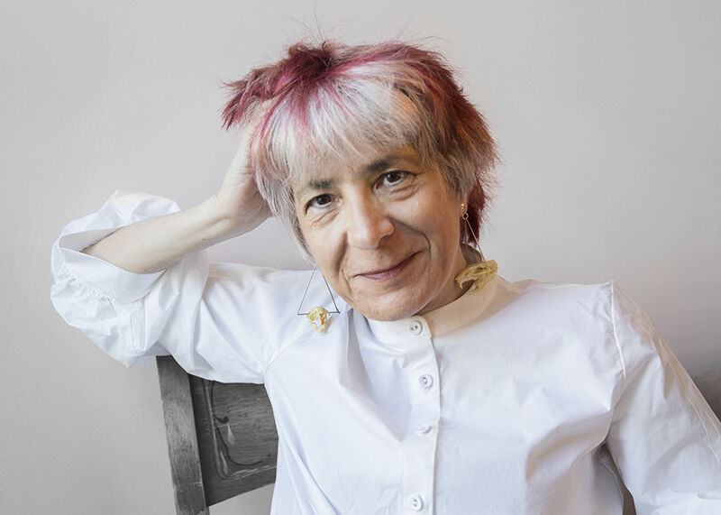 A white woman with short white hair with subtle lowlights, geometric earrings, and a crisp white collarless shirt poses seated, resting one arm on the back of her chair and smiling warmly.