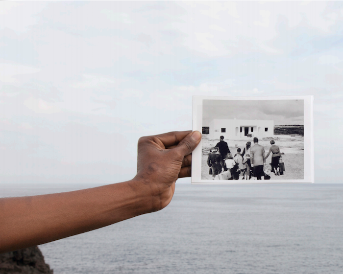 A hand holds up a black and white photo of a family with suitcases arriving at a white house before a vista of open water